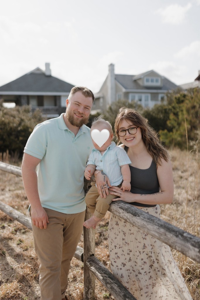 A family of three stands together outdoors near a wooden fence, with a house in the background. The parents smile while holding their toddler, who is partially obscured. The mother wears a casual top and a flowy skirt, while the father is dressed in a light shirt and trousers.
