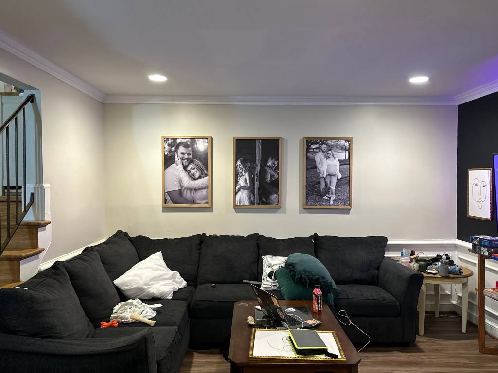 A cozy living room featuring a dark sectional sofa with colorful pillows and a cluttered coffee table, adorned with family photos in black and white on the walls.