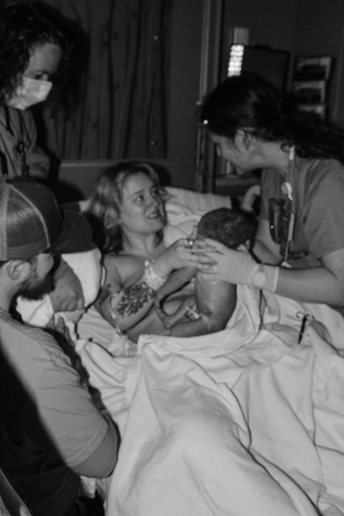 A black and white photograph of a mother in a hospital bed joyfully holding her newborn baby, surrounded by supportive family members and medical staff. The scene captures an emotional moment of connection and love.