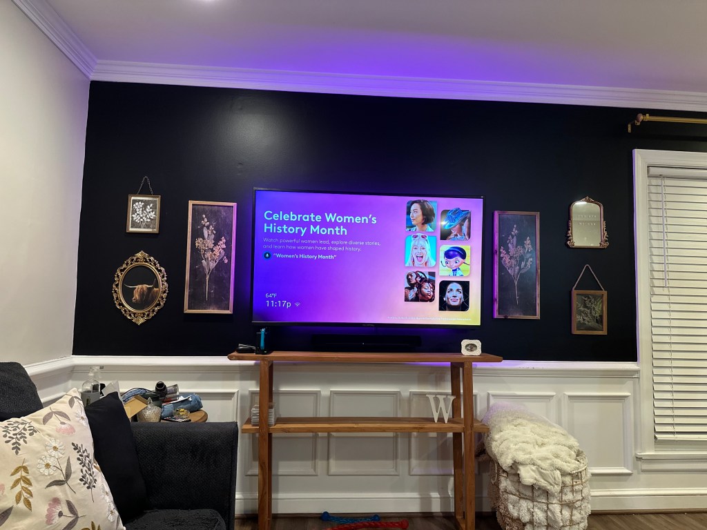 A living room wall featuring a black background, a TV displaying a slide that reads 'Celebrate Women's History Month', and several framed artworks and mirrors hung on the wall.
